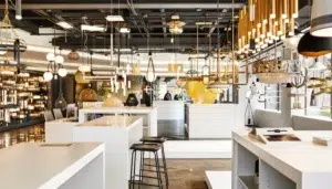 A modern lighting showroom featuring various hanging fixtures and lamps, with white display counters and a black stool in the foreground.