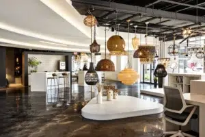 Spacious modern interior showroom with various pendant lights hanging. Display area features sleek counters, chairs, and decor on a reflective dark floor and open ceiling.