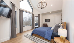 Modern bedroom with large windows, gray curtains, and a vaulted ceiling. Features include a blue bedspread, wall-mounted TV, round light fixture, and a small table with a lamp.