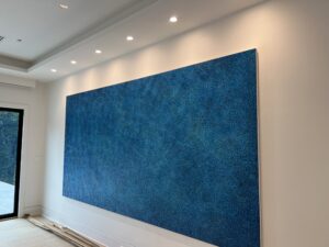 Large abstract painting with a blue, dot-like pattern hanging on a white wall under recessed lighting. A portion of a sliding glass door is visible on the left.