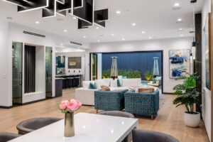 Modern open-plan living room with a white table and pink flowers in the foreground, blue and white seating, abstract art, and large glass doors opening to an outdoor area. Discover stylish lighting fixtures near me to complement this serene space.
