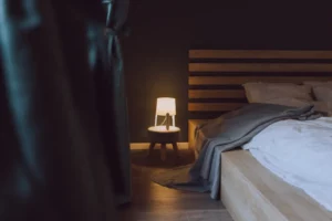 A wooden bed with white bedding and a gray blanket sits next to a small round nightstand with a lit lamp, creating cozy bedroom mood lighting in the dimly lit room.