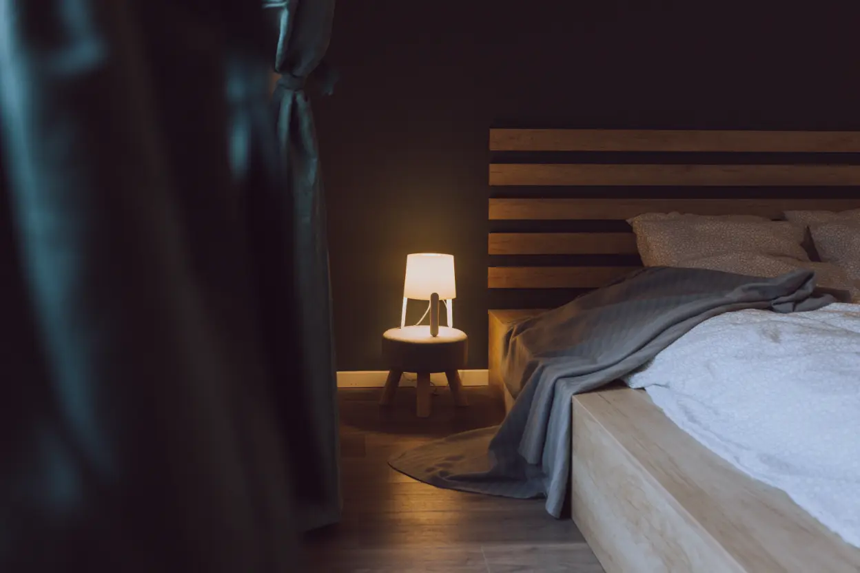 A wooden bed with white bedding and a gray blanket sits next to a small round nightstand with a lit lamp, creating cozy bedroom mood lighting in the dimly lit room.