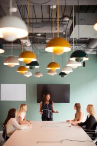 A woman stands and speaks to four seated women in a modern conference room with hanging lights and a wall-mounted TV.