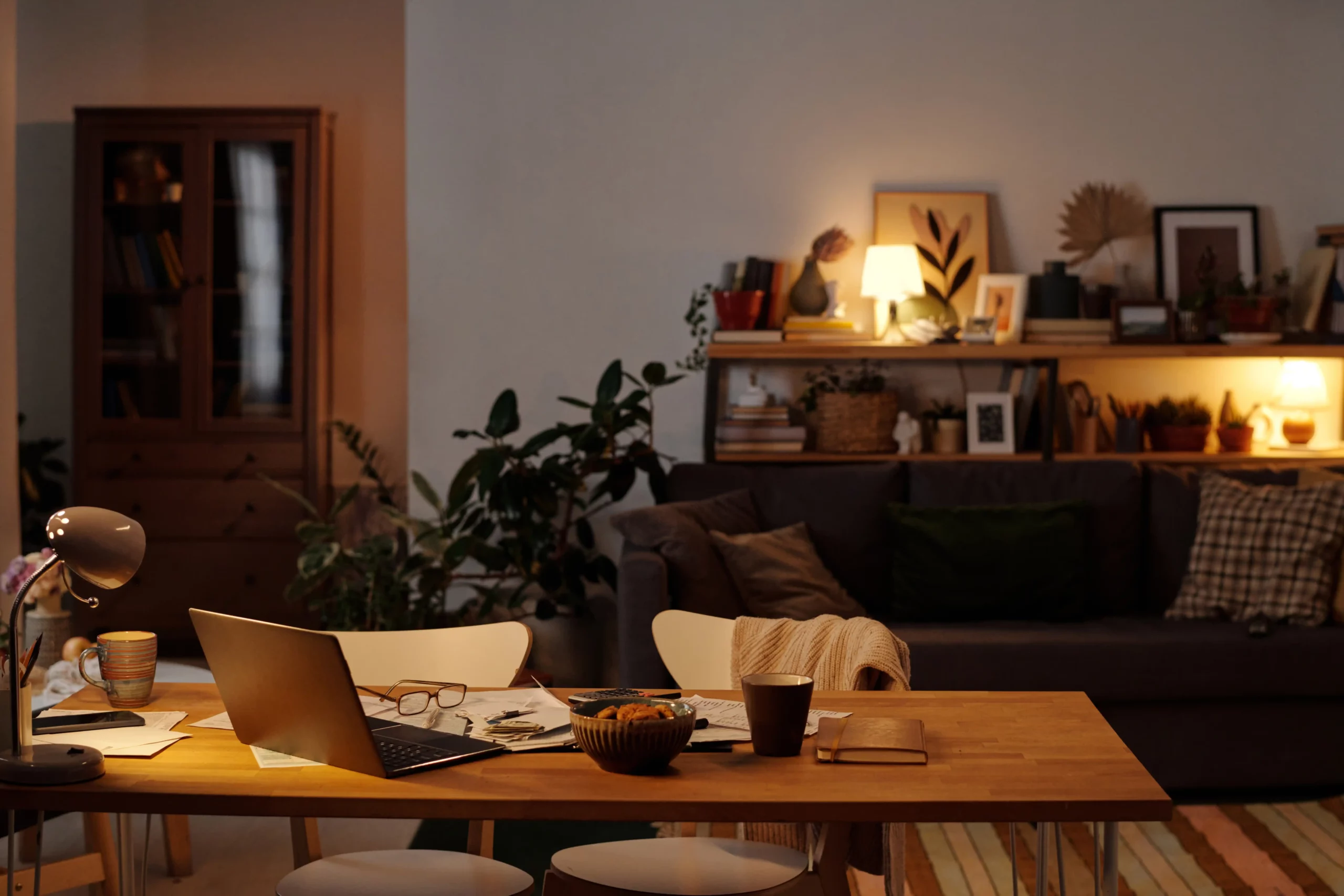 A wooden table with a laptop, glasses, papers, and snacks in a cozy living room with a sofa, plants, and bookshelves, showcasing ambient lighting for homes.